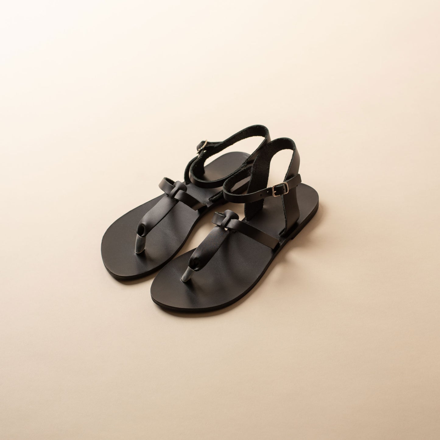 Pair of black designer flat sandals ankle strap on a beige background. They are flat sandals t strap.