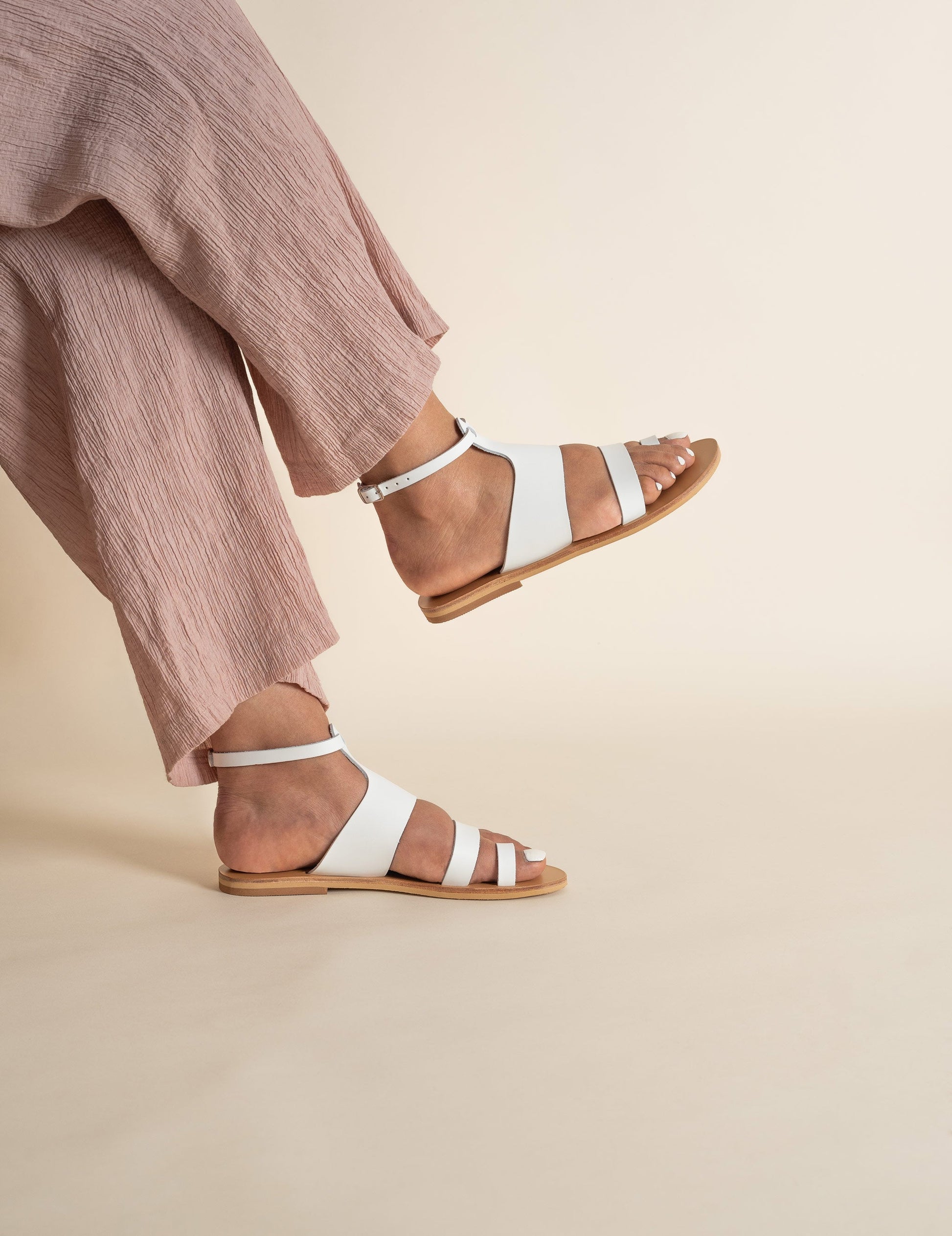 Woman wearing white flat sandals and pink pants with her legs crossed against a beige background. They are white ankle strap sandals.