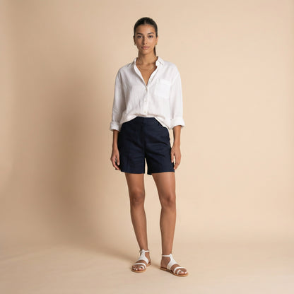 Woman wearing white flat sandals with blue shorts and a white shirt against a beige background. The sandals are white ankle strap sandals.