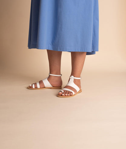Woman wearing white flat sandals and a blue dress against a beige background. The sandals are white ankle strap sandals.