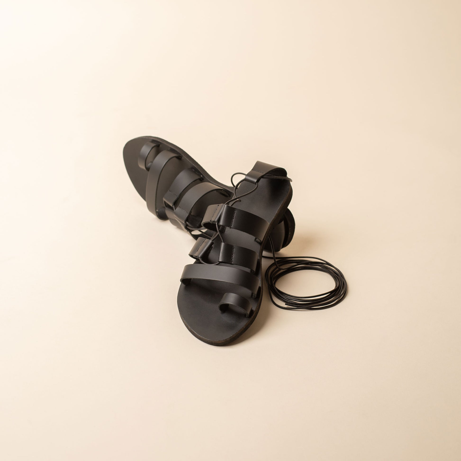 Black strappy flats shot against a beige background. They are flat sandals lace up.
