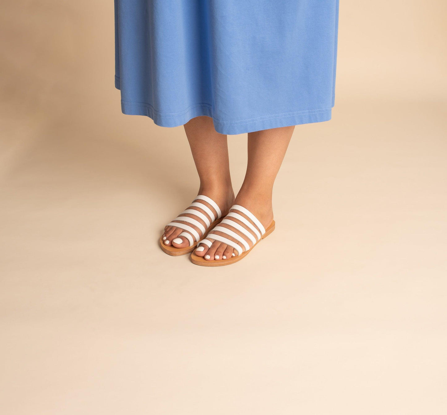 Woman wearing a pair of Harmonia white slide sandals and a blue dress against a beige background. These white leather sandals are white flats for women.