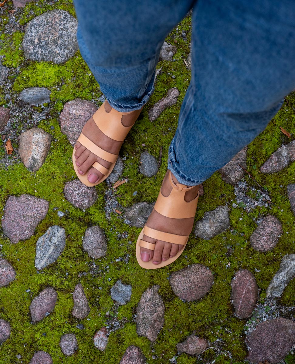 Top view of ĀRA flat sandals luxury brand in tan worn by a woman with blue jeans while standing on a stone floor that's covered in green moss. They are quality flat sandals.