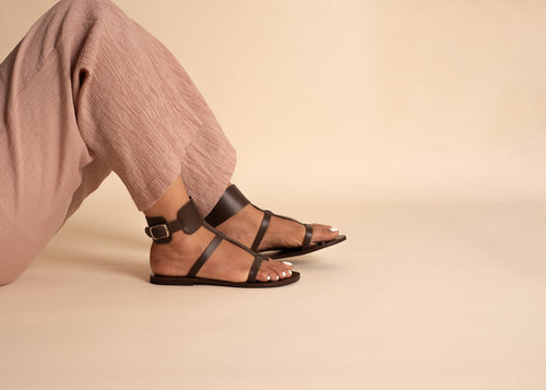 Woman wearing flat sandals luxury brand in chocolate brown with pink pants while sitting against a beige background. They are quality flat sandals.