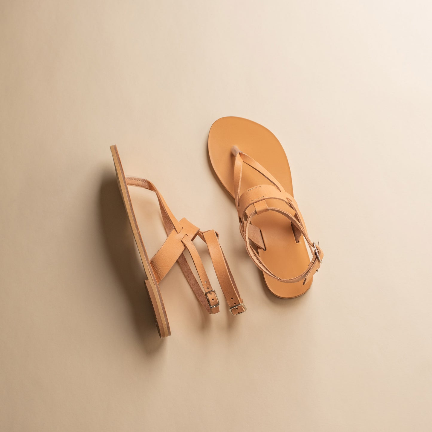 Top view of a pair of tan flat sandals designer on a beige background. They are women's flats with ankle straps.