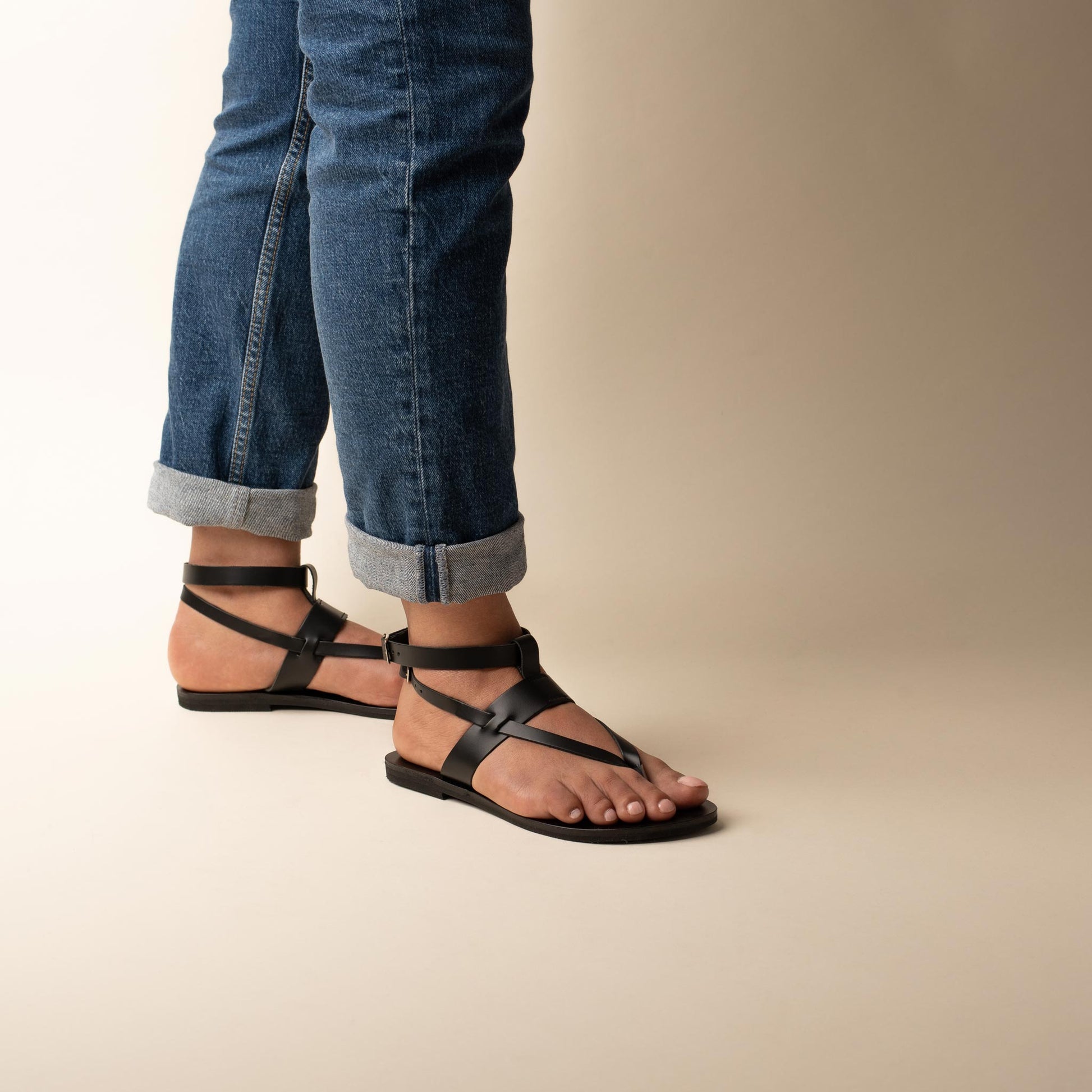 Woman wearing Rhea double dtrap sandals in black with blue cropped jeans against a beige background. They are black flats for women.