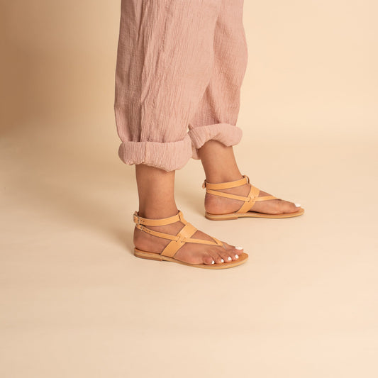 Woman wearing Rhea flat sandals designer in camel with pink cropped pants against a beige background. They are women's flats with ankle straps.