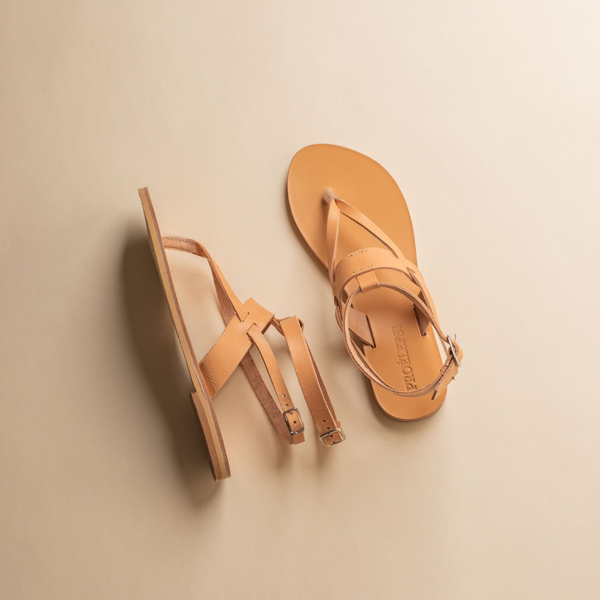 Top view of a pair of tan flat sandals designer on a beige background. They are women's flats with ankle straps.