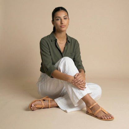 Woman wearing tan flat sandals t strap and a blue dress against a beige background. They are flat sandals beige.