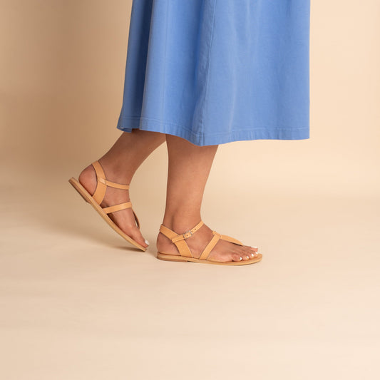 Woman wearing tan flat sandals t strap and a blue dress against a beige background. They are flat sandals beige.
